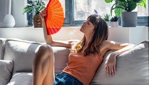 Eine Frau in kurzer Hose und Top sitzt auf einem Sofa und fächelt sich mit einem roten Fächer kühle Luft zu