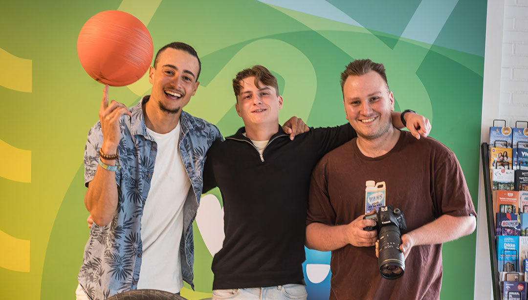 Drei junge Männer posieren für die Kamera. Einer hält eine Kamera in den Händen, der linke balanciert einen Basketball auf dem Zeigefinger. Der junge Mann in der Mitte hat die Arme um seine beiden Freunde gelegt.