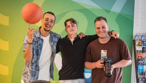 Drei junge Männer posieren für die Kamera. Einer hält eine Kamera in den Händen, der linke balanciert einen Basketball auf dem Zeigefinger. Der junge Mann in der Mitte hat die Arme um seine beiden Freunde gelegt.