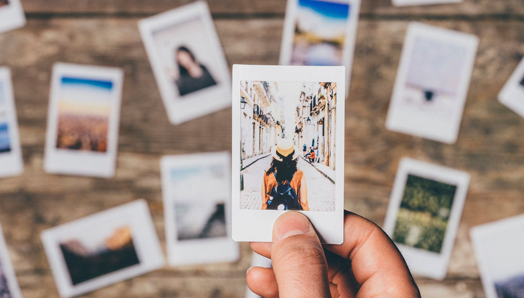 Eine Person hält ein Foto in der rechten Hand. Darauf ist eder Rücken einer Frau zusehen, die einen Hut und einen Rucksack trägt und durch eine Straße in einer Statd geht.
