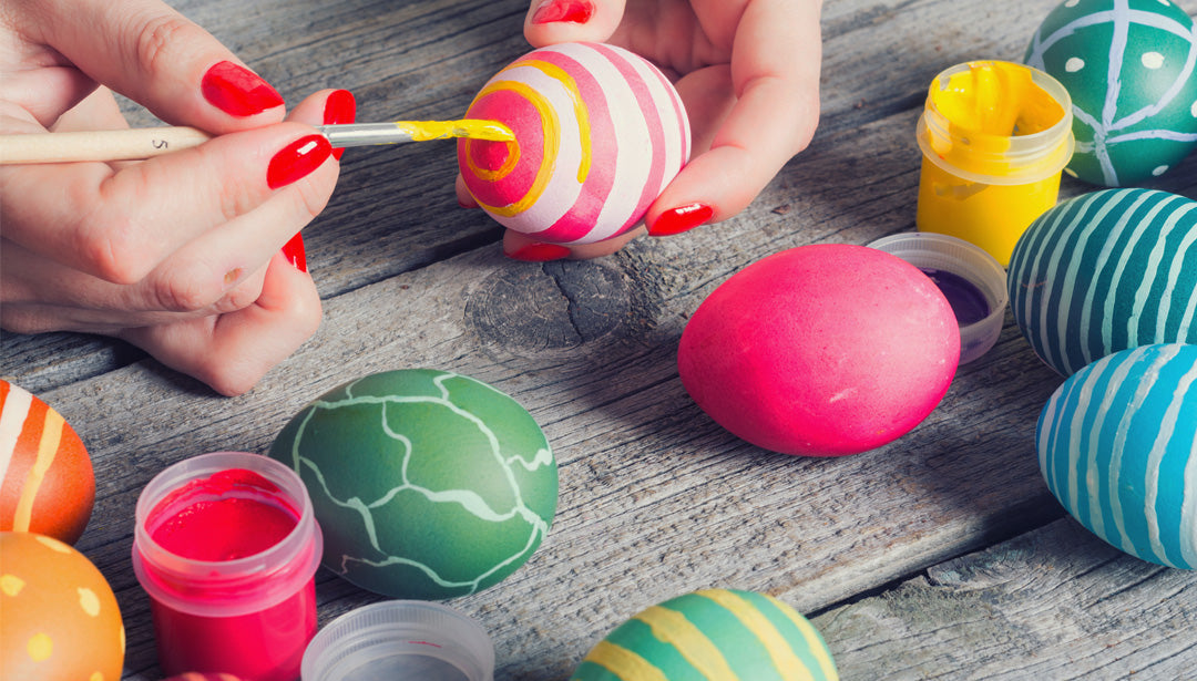 Eine Frau sitzt an einem Holztisch und bemalt Ostereier mit bunten Farben.