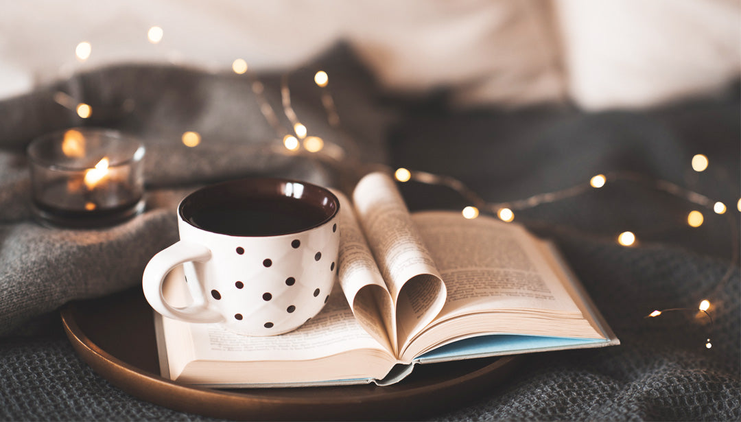 Eine gefüllte Tasse steht auf einem aufgeschlagenen Buch. Im Hintergrund sind eine kleine brennende Kerze und eine Lichterkette zu sehen. 