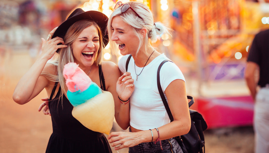 Zwei lachende, junge Frauen stehen vor einem Karussell auf einem Volksfest und essen Zuckerwatte. Es ist Sommer.